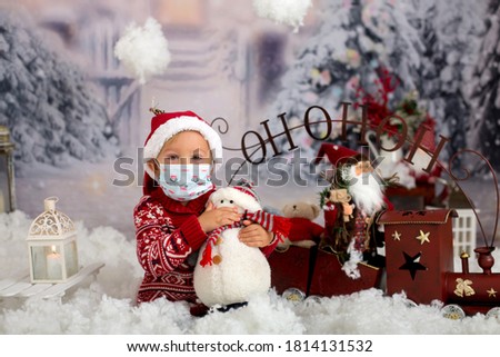 Cute boy, toddler child, having his christmas pictures taken playing in the snow outdoors, wearing protective mask, Christmas 2020 concept studio shot
