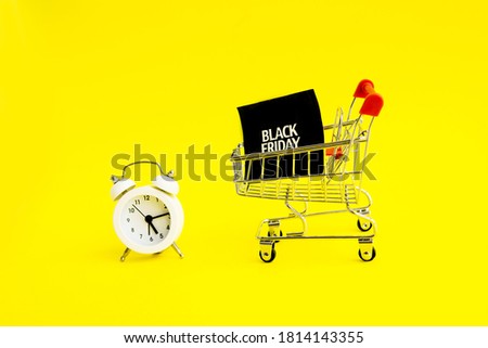 A white alarm clock, a shopping cart, and a black square that says black Friday on a yellow background. The topic of sales.