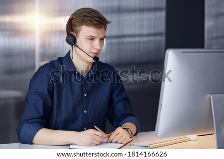 Young blond businessman using headset and computer in a darkened office, glare of light on the background. Startup business means working hard