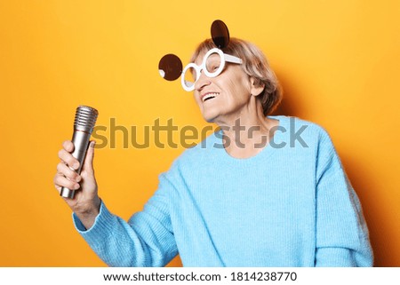 happy old woman with big eyeglasses holding a microphone and singing isolated on yellow background
