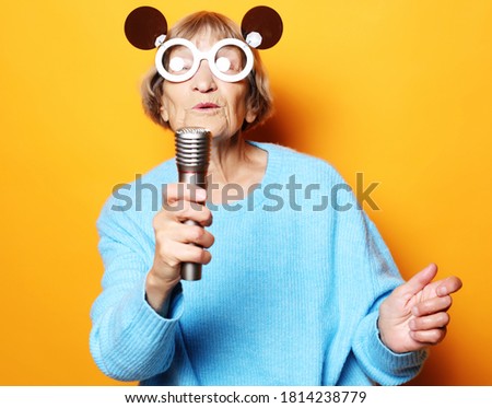 happy old woman with big eyeglasses holding a microphone and singing isolated on yellow background