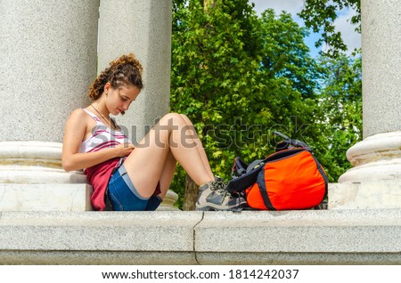 Stock photo of a young woman with her mobile smiling and adventurer. Curly girl sitting in marble building surrounded by adventure backpacks.
