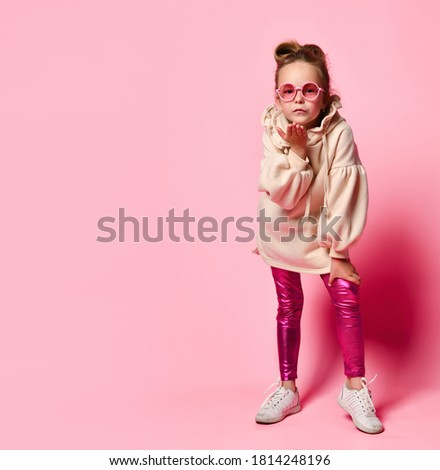 Serious little girl in stylish pink attire and round glasses leaning forward blowing a kiss to spectator. Full length shot isolated on pink, copy space. Children, gestures and emotions, snappy dresser