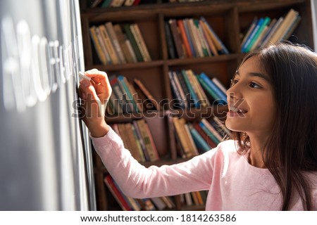 Happy indian kid primary school girl pupil holding chalk writing on blackboard. Smiling latin child preteen schoolgirl learning english alphabet letter handwriting standing in classroom. Close up view Royalty-Free Stock Photo #1814263586