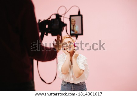 Female cameraman filming young woman posing with wireless headphones, using field monitor. Over pink background. Model has short dyed blond hair, wearing mom jeans and dress shirt. Focus on model.
