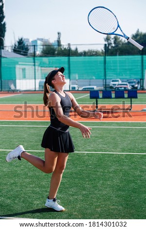 Teenage girl in a black sportive outfit training on a new tennis court, playing with the racket, flipping it in the air. Over a banner covering fence and high storey building. Side view.