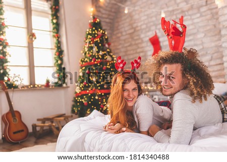 Couple in love wearing pajamas and deer antlers costume lying in bed on Christmas morning, having fun and enjoying their time together