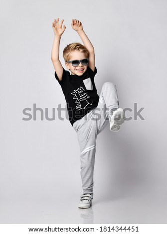 Playful frolic blond kid boy in sunglasses, black t-shirt with dinosaur print and gray pants stands holding hands and foot up over gray background