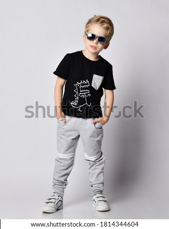 Cool blond kid boy in sunglasses, black t-shirt with dinosaur print and gray pants stands leaning sideways holding hands in pockets over gray background