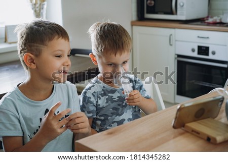 little boy making inhalation with nebuliser, his elder brother sitting next to him. children are watching cartoons on mobile phone