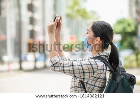 Young Asian beautiful woman travelers use her mobile phones to take the photo of view urban city downtown. She wearing protection mask for new normal safety travel trips.