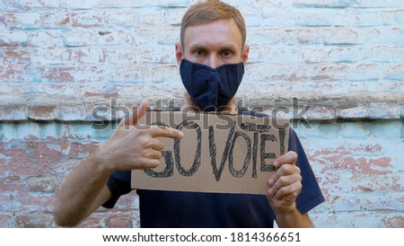 Man shows cardboard with Go Vote sign on brick wall urban background. Voting concept. Make the political choice, use your voice. Guy in protective face mask calls to go to the presidential elections.