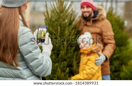 family, winter holidays and people concept - happy mother, father and little daughter buying christmas tree and taking picture with smartphone at street market