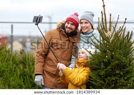 family, winter holidays and people concept - happy mother, father and little daughter buying christmas tree and taking picture with smartphone on selfie stick at street market