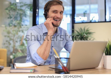 Cheerful young support phone male operator in headset, at workplace while using laptop, help service and client consulting call center concept