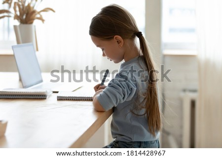 Side view focused small adorable caucasian girl sitting at desk with correct posture straight back, preparing homework alone indoors, smart little kid studying remotely, homeschooling concept. Royalty-Free Stock Photo #1814428967