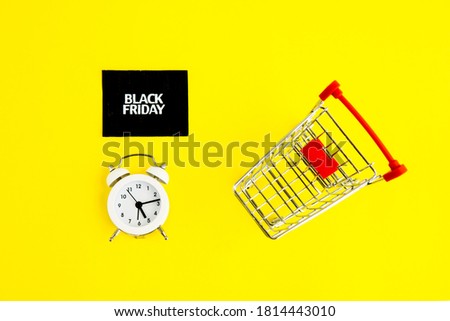 A white alarm clock, a shopping cart, and a black square that says black Friday on a yellow background. The topic of sales.