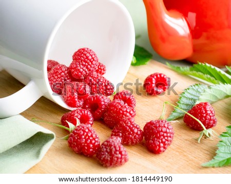 Ripe raspberry berries on a wooden background are poured out of a white cup. There's a kettle next to it. selective focus. wholesome dessert.