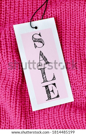 Word Sale on price tag on bright pink knitted wool fabric with folds or sweater. A gray textured cardboard label card attached to the black tape on the clothes. Vertically shopping concept.