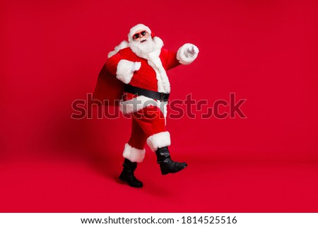 Full length photo of retired old man grey beard hold bag presents walk humming christmas song wear santa costume gloves coat belt sunglass headwear black boots isolated red color background