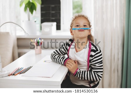 The girl is sitting at the table holding a blue felt-tip pen with her nose and lips. A girl plays before completing her homework. Home games, homework, home training, social distance