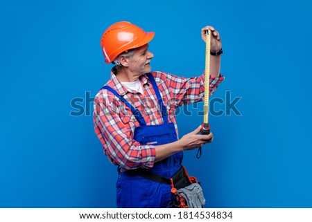 Professional picture of  senior man builder and plumber in orange helmet ready for a work holding centimeter isolated on blue background.
