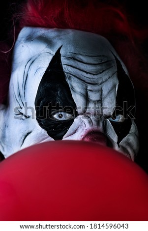 closeup of a scary evil clown with red hair and white eyes, staring at the observer, with a red balloon in front of him, against a black background