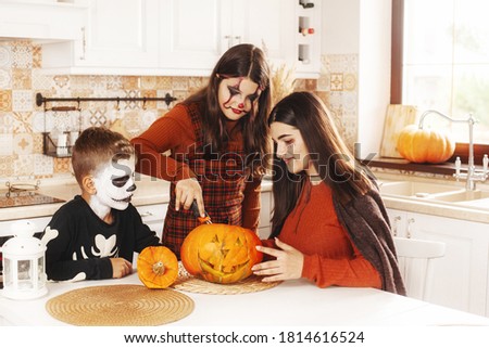 Sisters and brother at home in the kitchen in costumes and makeup for Halloween, carving a pumpkin, playing and laughing.