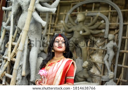 Portrait of beautiful Indian Bengali female woman in red and white traditional ethnic saree and jewellery in front of the clay idol of Hindu goddess Durga. Indian culture, religion and fashion