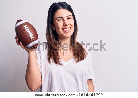 Young beautiful brunette woman playing rubgy holding football ball over white background looking positive and happy standing and smiling with a confident smile showing teeth