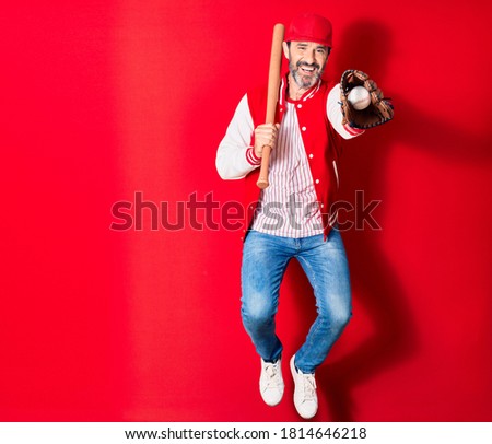 Middle age handsome man wearing sporty clothes smiling happy. Jumping with smile on face playing baseball using bat ,ball and glove over isolated red background
