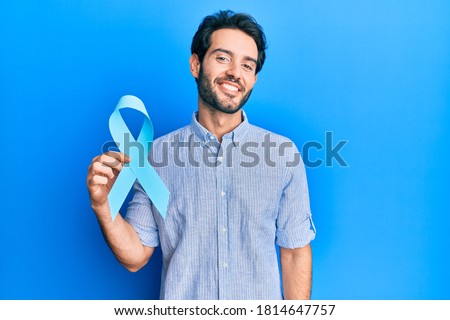 Young hispanic man holding blue ribbon looking positive and happy standing and smiling with a confident smile showing teeth  Royalty-Free Stock Photo #1814647757
