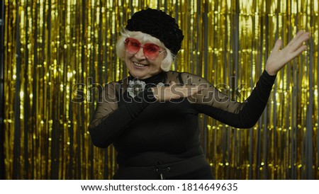 Senior old woman in red sunglasses dances, celebrates, listens music. Energetically moving to the rhythm, relaxing, enjoying, having fun, smiling. Elderly stylish lady grandma in studio