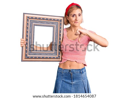 Young blonde woman holding empty frame with angry face, negative sign showing dislike with thumbs down, rejection concept 