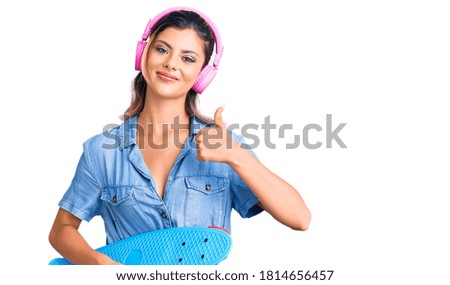 Young beautiful woman wearing headphones and holding skate smiling happy and positive, thumb up doing excellent and approval sign 