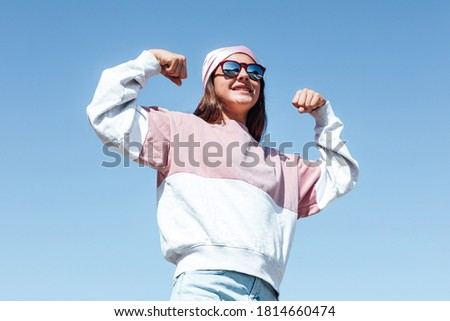 Girl woman with sunglasses and pink head scarf on her head, squeezes her arm as a sign of strength, on October 19,  International Breast Cancer Day, with the sky like background. Breast cancer concept