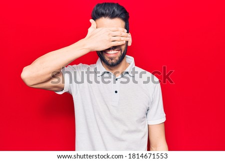 Young handsome man with beard wearing casual polo smiling and laughing with hand on face covering eyes for surprise. blind concept. 