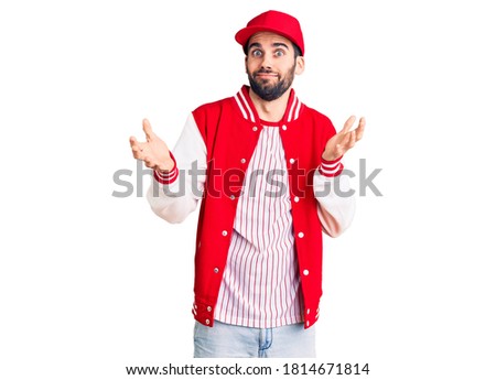 Young handsome man with beard wearing baseball jacket and cap clueless and confused expression with arms and hands raised. doubt concept. 