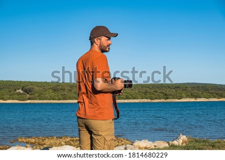 Photographer man in t-shirt and cargo pants walking around sea shore with DSLR camera and shooting nature, half body portrait, landscape photography concept
