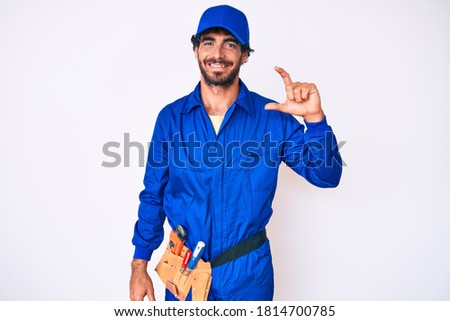 Handsome young man with curly hair and bear weaing handyman uniform smiling and confident gesturing with hand doing small size sign with fingers looking and the camera. measure concept. 