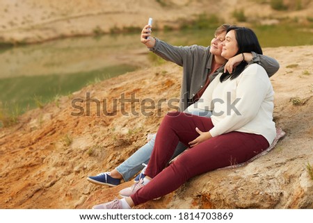 Lesbian couple making selfie portrait on mobile phone while sitting on the hill on the nature