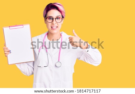Young beautiful woman with pink hair wearing doctor stethoscope holding clipboard with medical report smiling happy and positive, thumb up doing excellent and approval sign 