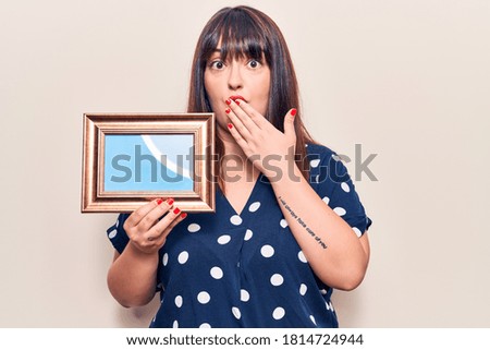 Young plus size woman holding empty frame covering mouth with hand, shocked and afraid for mistake. surprised expression 