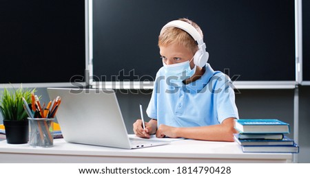 Small Caucasian teen boy in medical mask and headphones sitting at desk in school. Teenage schoolboy doing homework and writing exercise in copybook in front of laptop computer. Covid-19 concept.