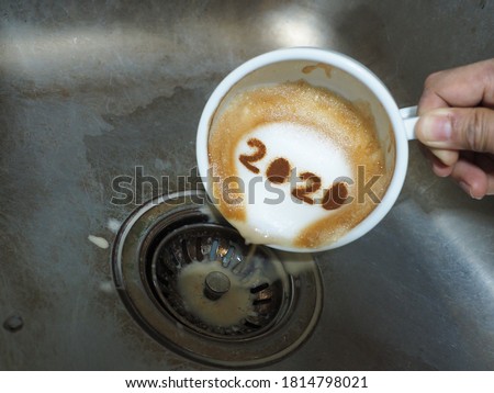 Goodbye 2020, a year that many want time to flies by. Holidays food art theme hand holding and pouring coffee with number 2020 on frothy surface from white cup over dirty stainless steel kitchen sink. Royalty-Free Stock Photo #1814798021