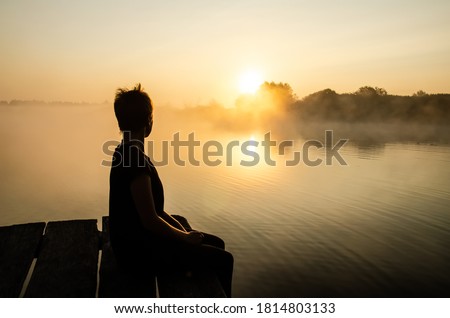 Silhouette of a person sitting on the pier against the sunset sky. Loneliness concept. The female looking at the rising sun over the lake in the fog. Reflection of the sun in the water surface. Royalty-Free Stock Photo #1814803133