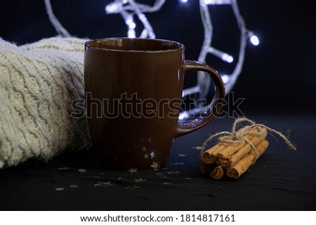 A brown cup of coffee, next to cinnamon sticks and star anise. Knitted background and Christmas lights.