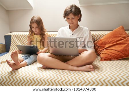 Little children, cute brother and sister sitting on sofa and using laptop and digital tablet, watching cartoons or playing games while spending time together at home