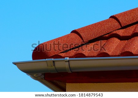 gray silver shallow zink eavestrough or gutter. red brown color sandy textured modern concrete roof tile closeup detail with half round ridge tiles.  house construction concept. clear blue sky Royalty-Free Stock Photo #1814879543