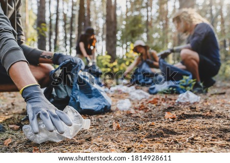 Close up of hands with plastic bottle with group of young people friends volunteers collecting plastic bottles to trash bags in forest background. Ecology concept Royalty-Free Stock Photo #1814928611